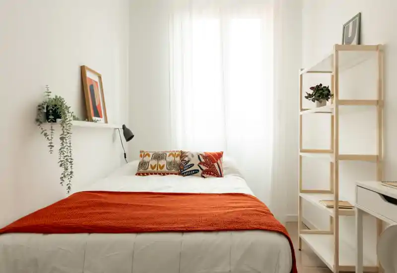 Camera da letto luminosa e minimal con letto ben rifatto, copriletto arancione caldo e cuscini decorativi; la luce naturale attraverso le tende crea un'atmosfera tranquilla e accogliente.