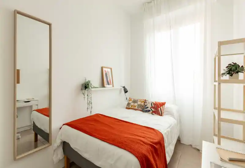 Camera da letto luminosa e minimal con letto matrimoniale in lenzuola bianche e un caldo plaid arancione; tende leggere diffondono la luce naturale creando un'atmosfera ariosa.