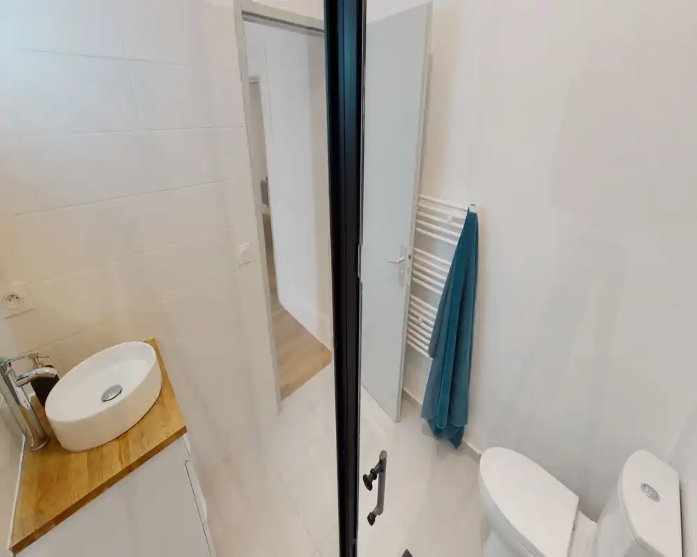 Salle de bain moderne et lumineuse avec toilette blanche, sèche-serviettes mural et lavabo posé sur un plan en bois ; carrelage clair et lignes épurées pour un style minimal.