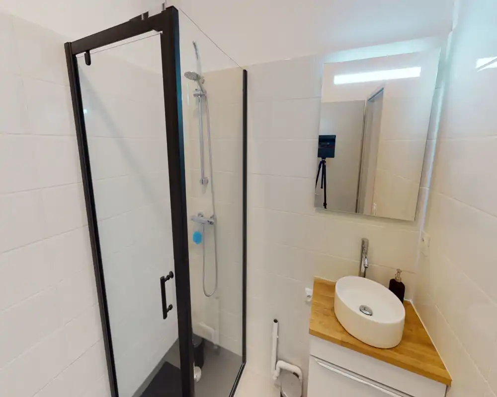 Salle de bains moderne et compacte avec douche vitrée, robinet mural et vasque ronde posée sur une coiffeuse en bois. Carrelage blanc et grand miroir pour une atmosphère propre et lumineuse.