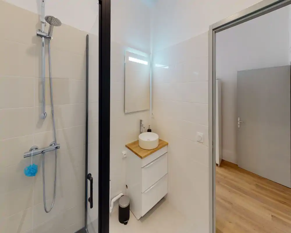Salle de bain moderne et compacte avec cabine de douche vitrée, douche à effet pluie murale et petit meuble vasque rond avec miroir ; finitions propres et lumineuses, parquet visible à l'extérieur.