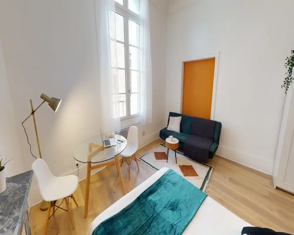 Chambre lumineuse et moderne avec le lit bien visible au premier plan, un petit coin salon avec un canapé vert et un bureau en verre près de la fenêtre — idéale pour un séjour en ville.