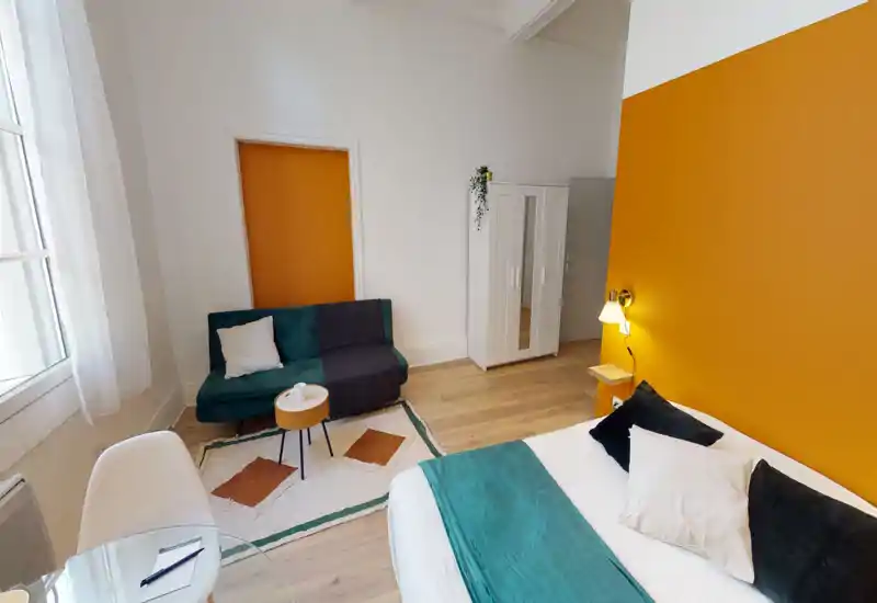 Chambre lumineuse et moderne avec grand lit, jeté de lit et coussins bleu-vert, mur d'accent orange et coin salon avec canapé et table basse.