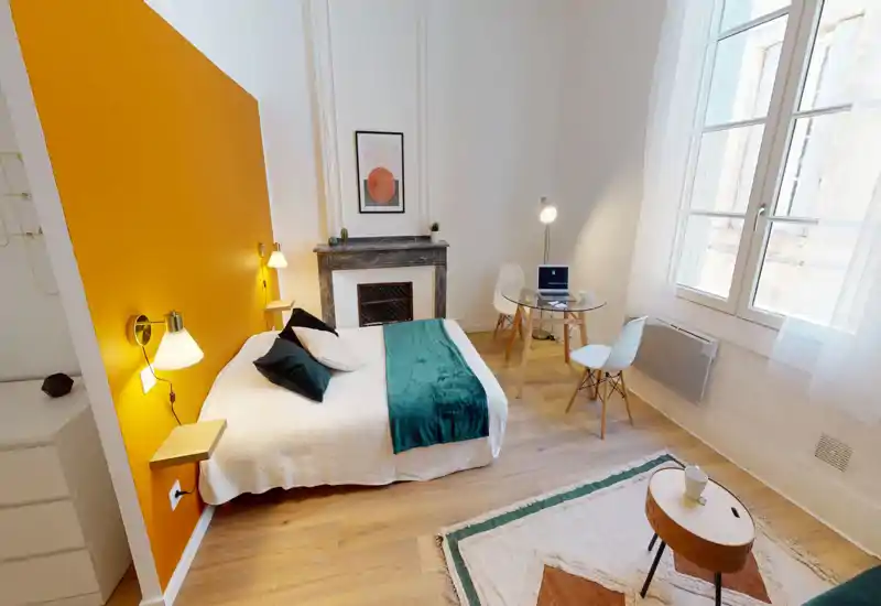 Chambre lumineuse et élégante avec un lit complet contre un mur d'accent moutarde, lampes de chevet, une cheminée et une petite table repas/travail près d'une grande fenêtre baignée de lumière naturelle.