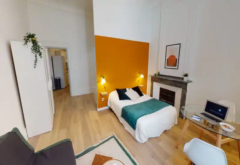 Chambre lumineuse et décorée avec un lit double contre un mur ocre, lampes de chevet et accessoires soignés pour une ambiance chaleureuse.