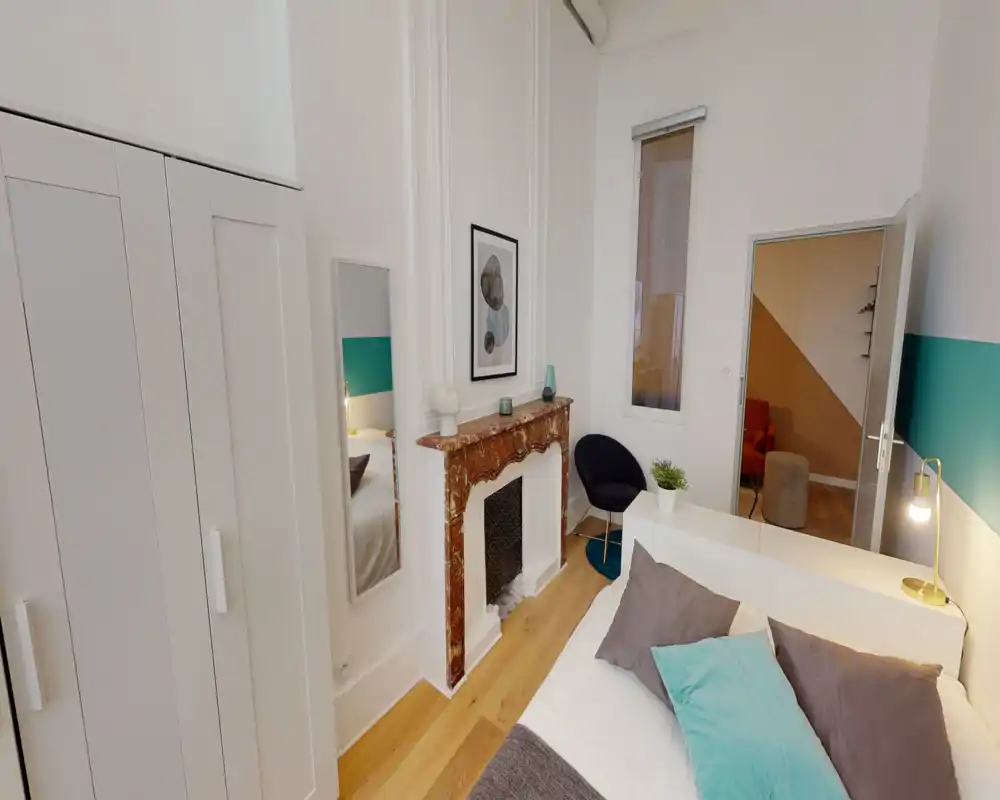Chambre moderne et lumineuse avec un lit double au premier plan, coussins décoratifs et une cheminée en marbre. Murs blancs, parquet, armoire et fauteuil cosy.