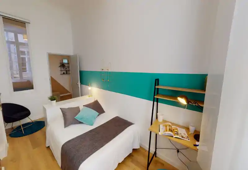 Chambre moderne et lumineuse avec un lit double complet, coussins décoratifs et un petit espace de travail. Murs blancs avec une bande turquoise, sol en bois et textiles doux qui rendent la pièce chaleureuse.
