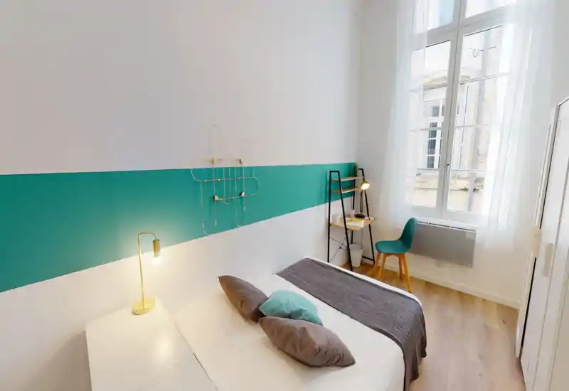 Chambre moderne et lumineuse avec un lit double bien fait, coussins décoratifs et une bande turquoise sur le mur; grande fenêtre apportant une belle lumière naturelle et un coin bureau agréable.