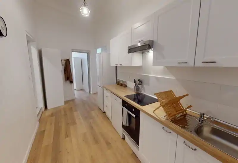 Cuisine moderne en enfilade, lumineuse, avec meubles blancs, plans de travail en bois, four encastré et évier; propre et bien éclairée, idéale pour un appartement compact.