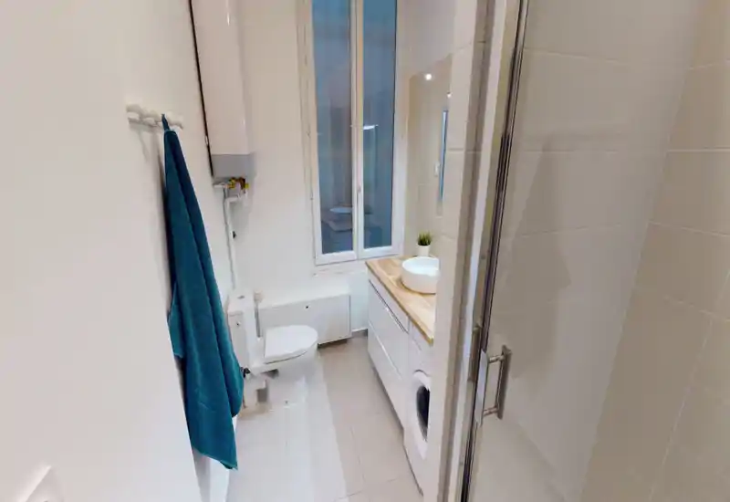 Salle de bain compacte et lumineuse avec WC, vasque sur plan en bois, machine à laver sous le plan et porte de douche en verre. Carrelage blanc et serviette bleue pour une atmosphère moderne et fraîche.