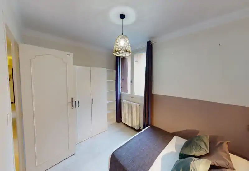 Chambre lumineuse au style épuré avec un lit double habillé de linge neutre, coussins et plaid; armoire et étagères près de la fenêtre offrant rangement et lumière naturelle.