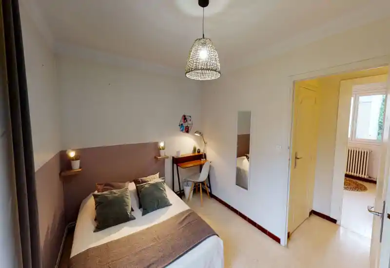 Chambre lumineuse et bien aménagée avec un lit double, coussins décoratifs, petit bureau et suspension lumineuse — chaleureuse et accueillante.