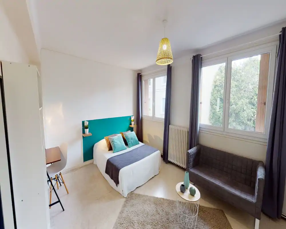 Bright, well-styled bedroom with a double bed against a teal headboard, large windows providing natural light, and a small sitting area with a sofa and coffee table.