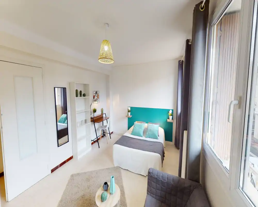 Bright, well-styled bedroom with a double bed, teal headboard and decorative cushions, a small desk and shelving. Large window provides natural light and a cozy seating area with rug and coffee table.