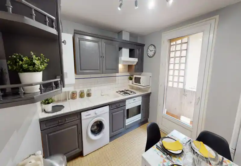 Cuisine lumineuse et bien équipée avec meubles gris, four encastré et plaque à gaz, machine à laver et petite table près d'une porte vitrée qui apporte la lumière naturelle.