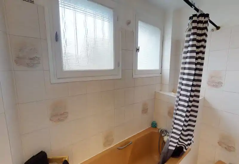 Salle de bains compacte avec baignoire, rideau de douche rayé et fenêtres dépoli apportant une lumière naturelle agréable ; carrelage décoré.