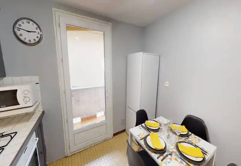 Cuisine compacte avec une petite table dressée pour quatre, un grand réfrigérateur blanc, un micro-ondes et une porte vitrée donnant sur un balcon qui apporte la lumière naturelle.