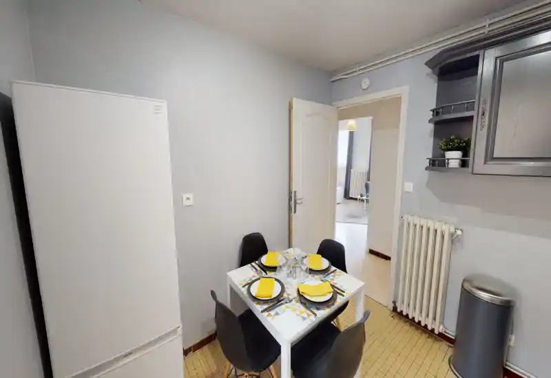 Coin cuisine/salle à manger compact avec une petite table pour quatre, un grand réfrigérateur et des placards muraux ; lumineux, propre et mis en scène pour un repas convivial.