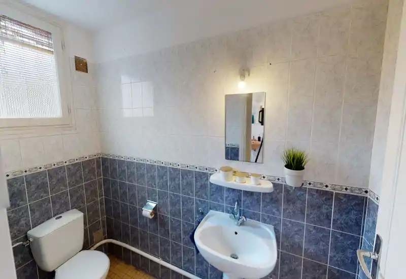 Salle de bains carrelée compacte avec lavabo, miroir, WC et petite fenêtre ; propre et lumineuse.