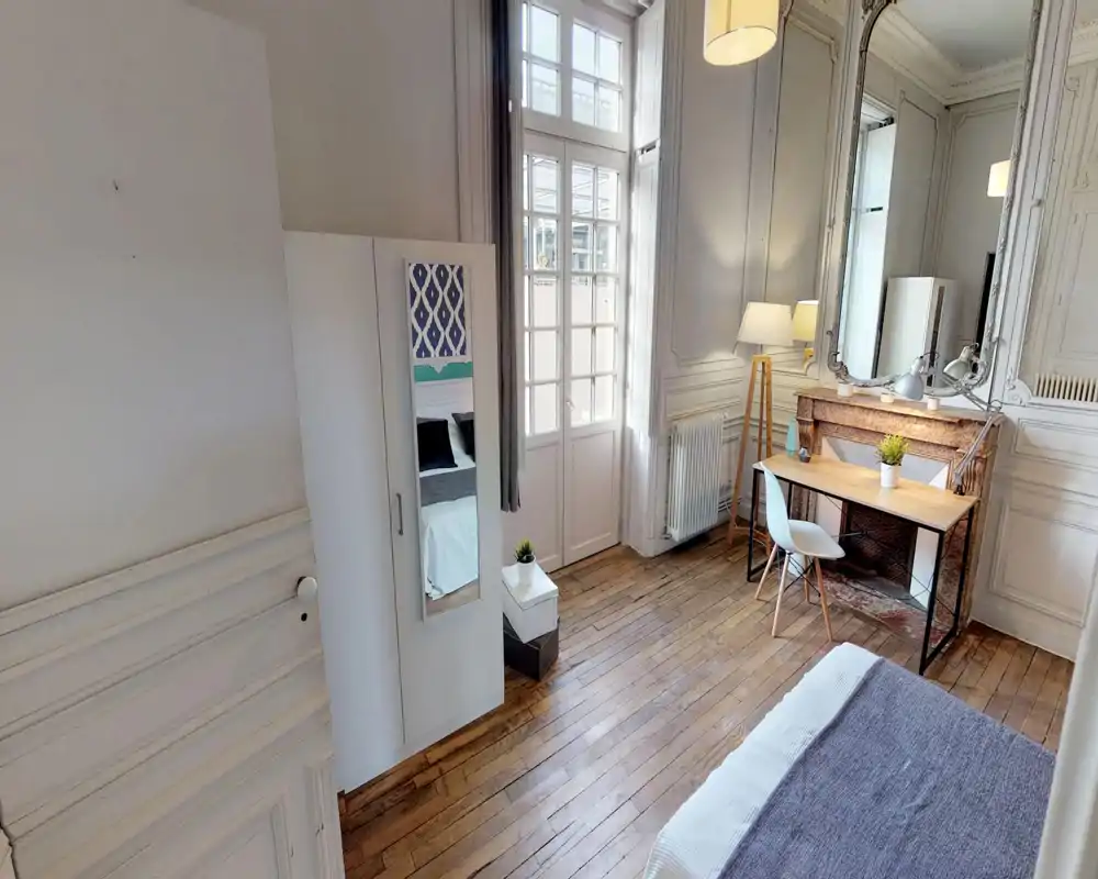 Bright, elegant bedroom with wooden floors, large windows and a cozy desk area; bed partially visible at the corner and mirrored in the wardrobe door.