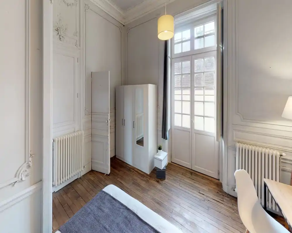 Bright, elegant bedroom with high ceilings, decorative mouldings and a visible bed in the foreground; tall French windows let in soft natural light and a white wardrobe with mirror completes the layout.