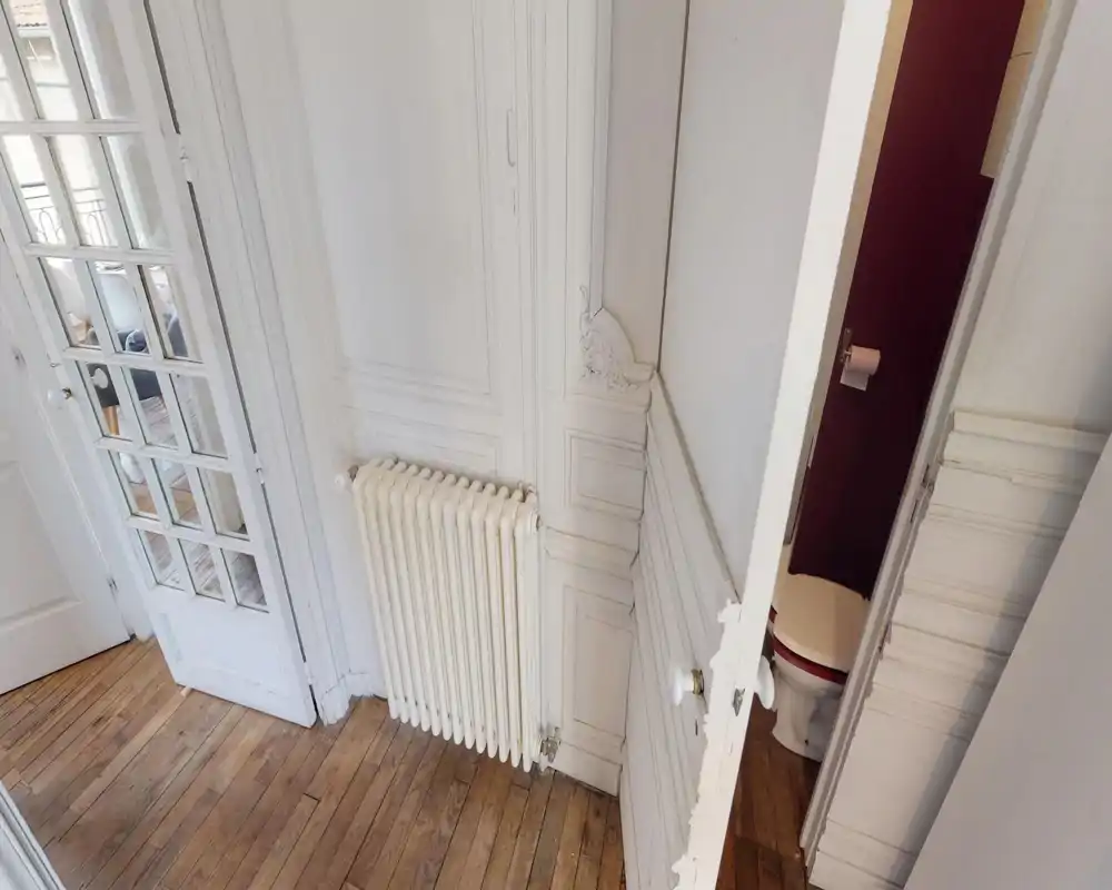 Small entry/hallway area with decorative wall moulding, a radiator and a partially open door leading to a small WC. Wooden floors and a glazed door to an adjacent room are visible.