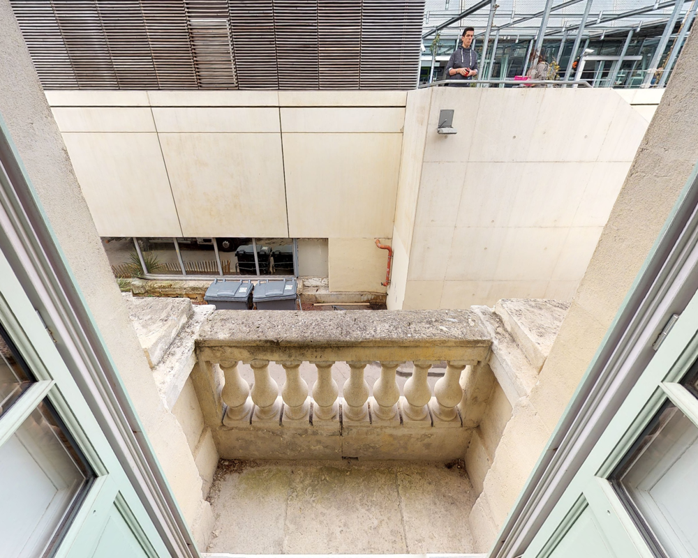 Small stone balcony overlooking a narrow urban lightwell; classical balustrade and worn floor visible, framed by open windows.