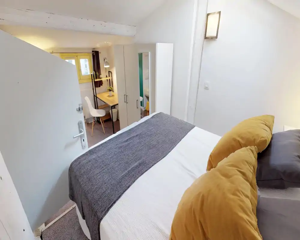 Bright, cozy bedroom with a neatly made double bed, mustard pillows and a grey runner; wardrobe and a small desk with chair near a window create a practical, well-lit space.