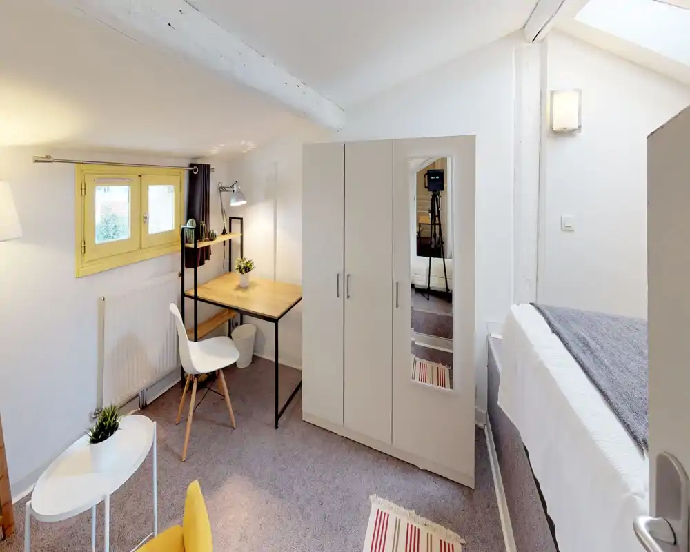 Cozy attic bedroom with a partial bed view, a compact wardrobe with mirror, a wooden desk and a bright skylight. Ideal for a single occupant seeking a well-lit work-and-sleep nook.