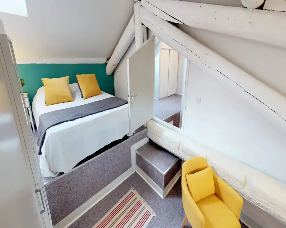 Bright attic bedroom with exposed painted beams, a raised double bed dressed in white linens and yellow accent pillows, and a cozy yellow armchair — stylish and inviting.