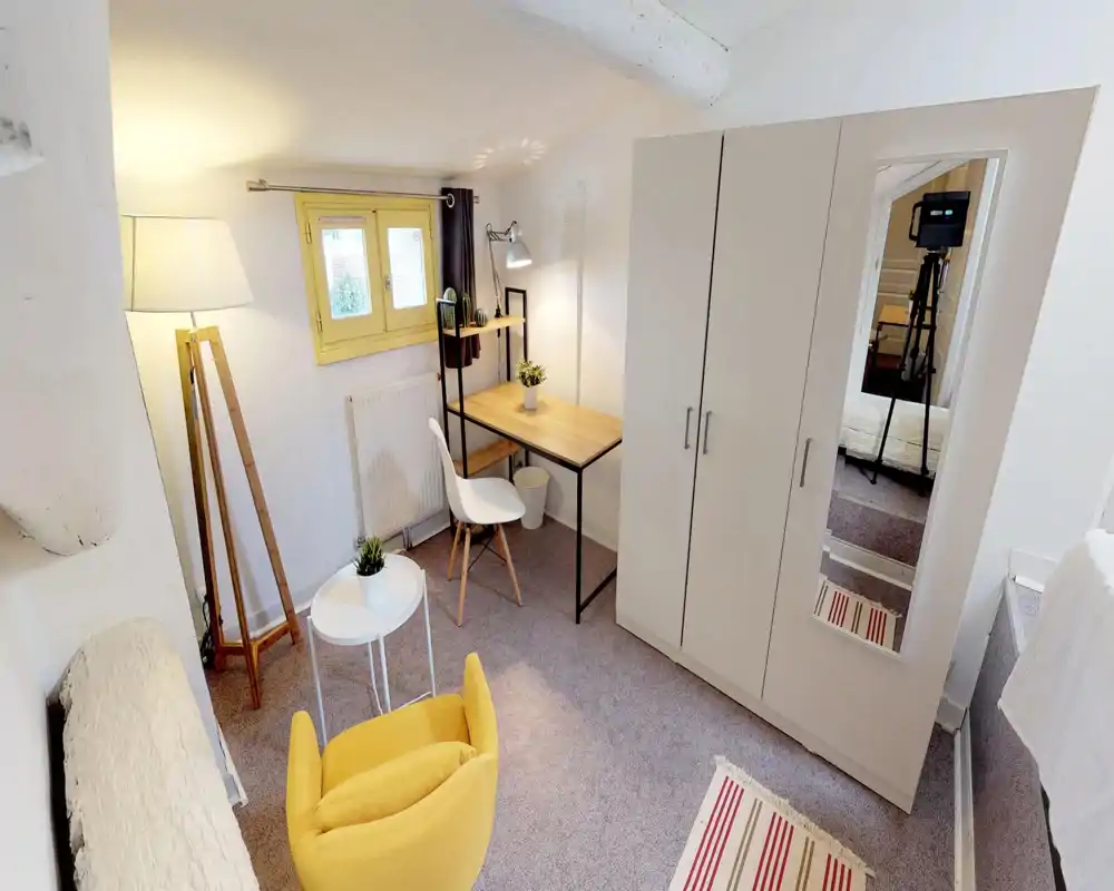 Bright, compact bedroom corner with a small workspace and wardrobe. The room features a cozy yellow armchair, wooden desk with shelving and a full-height wardrobe with mirror reflecting the bed.