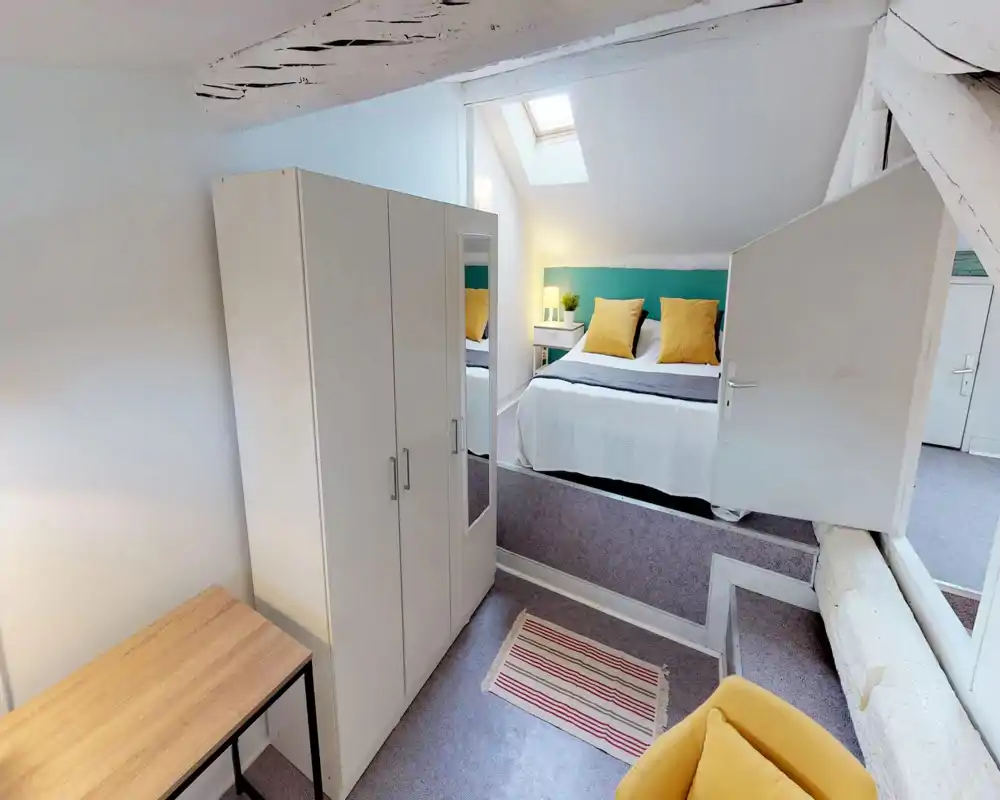 Bright, well-staged attic bedroom with a skylight, teal headboard and yellow cushions; includes wardrobe, bedside table and a comfortable double bed.
