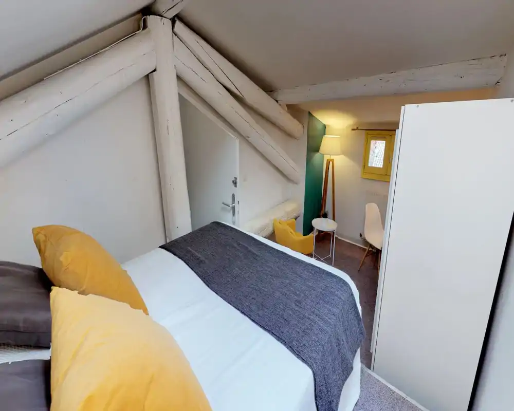 Charming attic bedroom with exposed white beams, a neatly made double bed with yellow accent pillows and a grey runner, and a cozy seating corner lit by a floor lamp.