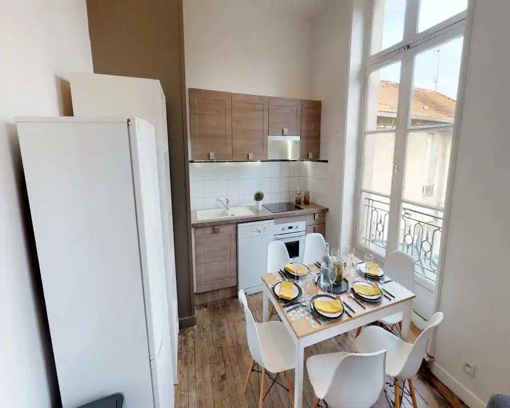 Bright, compact kitchen with wood cabinets, integrated oven and dishwasher, and a set dining table by a large window that fills the room with natural light.