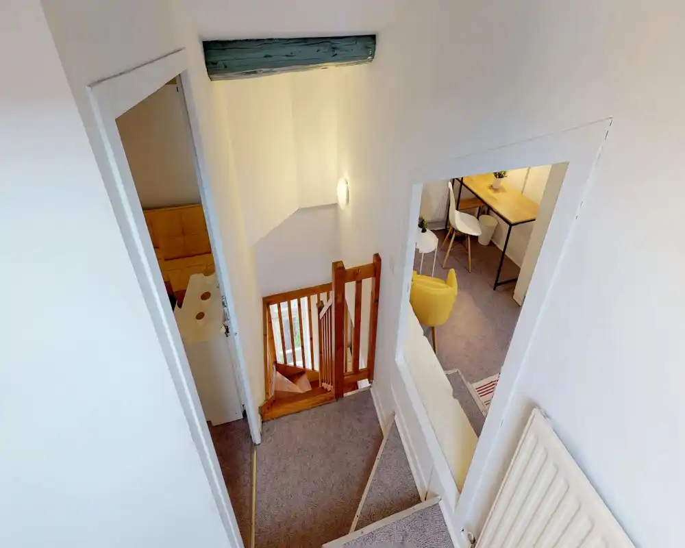 Small upstairs landing with carpeted floor, wooden stair gate and warm wall light; visible views into adjacent rooms with a yellow chair and a desk.