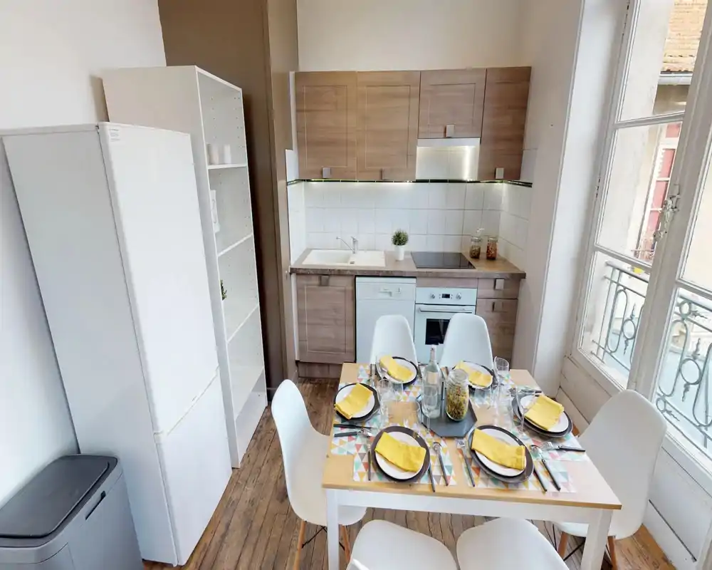 Compact, modern kitchenette with wood-effect cabinets, built-in oven and induction hob. A small dining table set for four sits in the foreground next to a large window that brings abundant natural light.