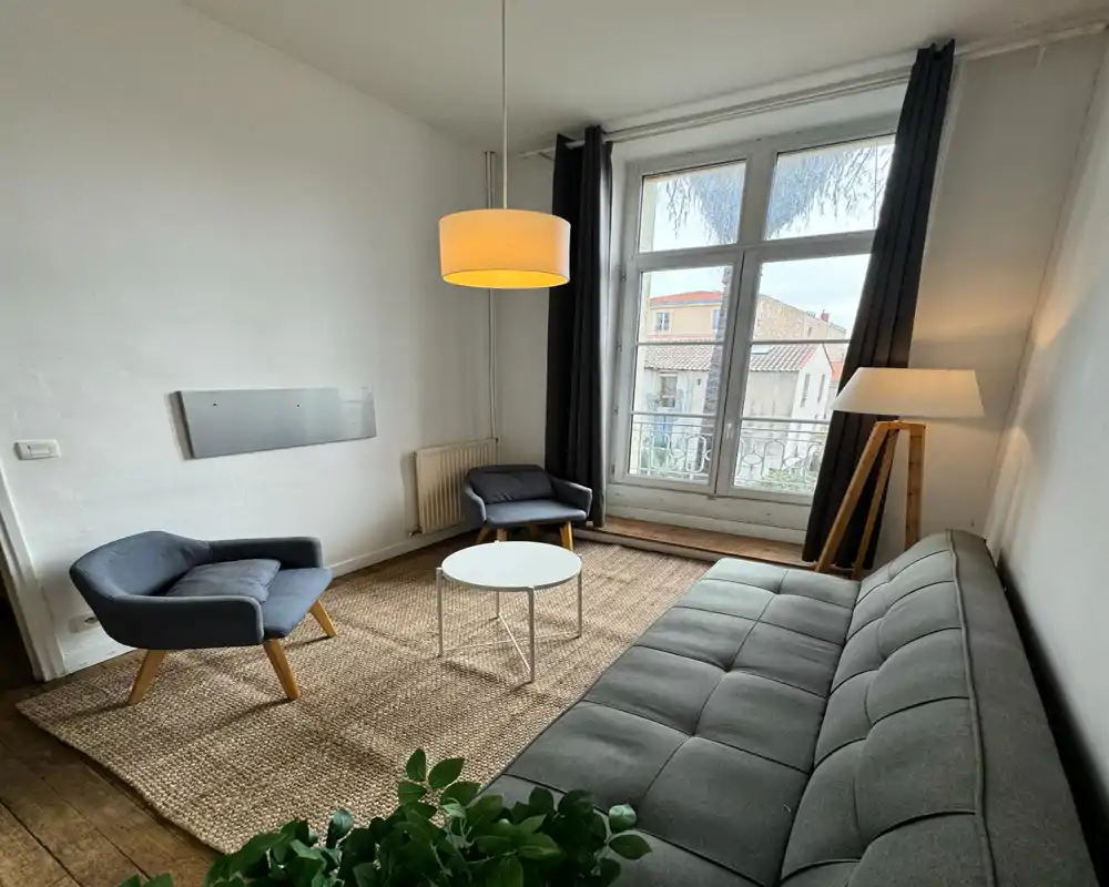 Cozy, well-lit living area with a grey sofa, two armchairs, a small round coffee table and large window providing natural light.