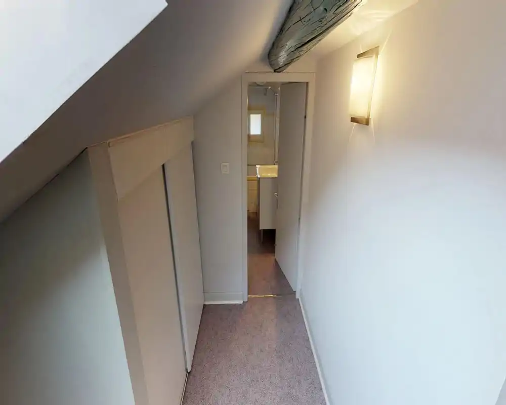 Narrow upstairs hallway/landing with sloped ceiling, built-in storage doors on the left and a small bathroom visible through an open door at the end. Soft wall sconce provides warm light; suitable for describing a compact access corridor.