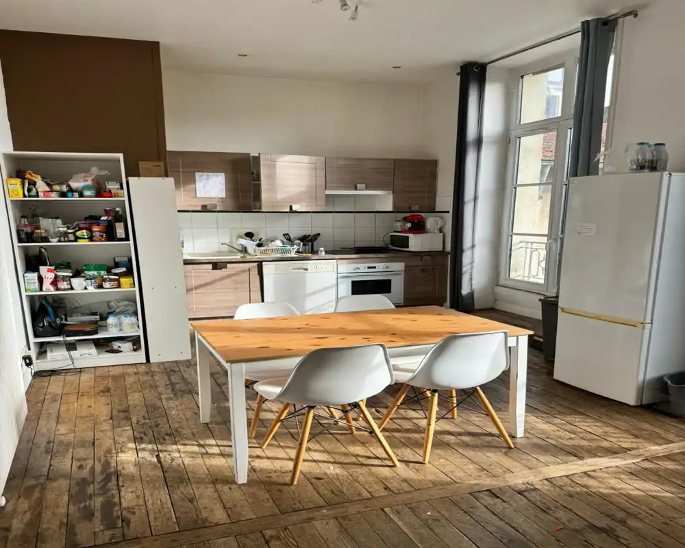 Sunny fitted kitchen with wooden floor and a central dining table for four; modern cabinets, dishwasher and tall fridge visible, ideal for shared living.