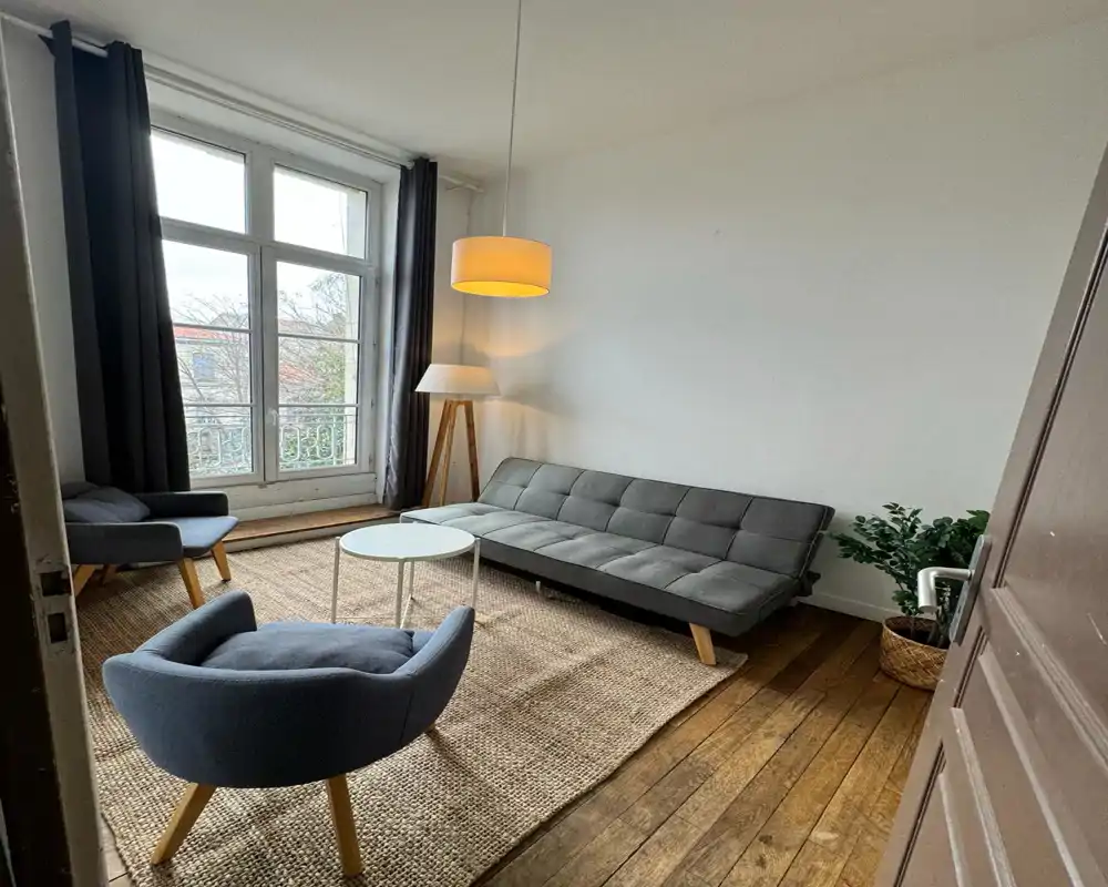 Bright, cozy living room with a gray sofa, two armchairs, a round coffee table and a large window letting in natural light; neutral tones and textured rug create a welcoming atmosphere.