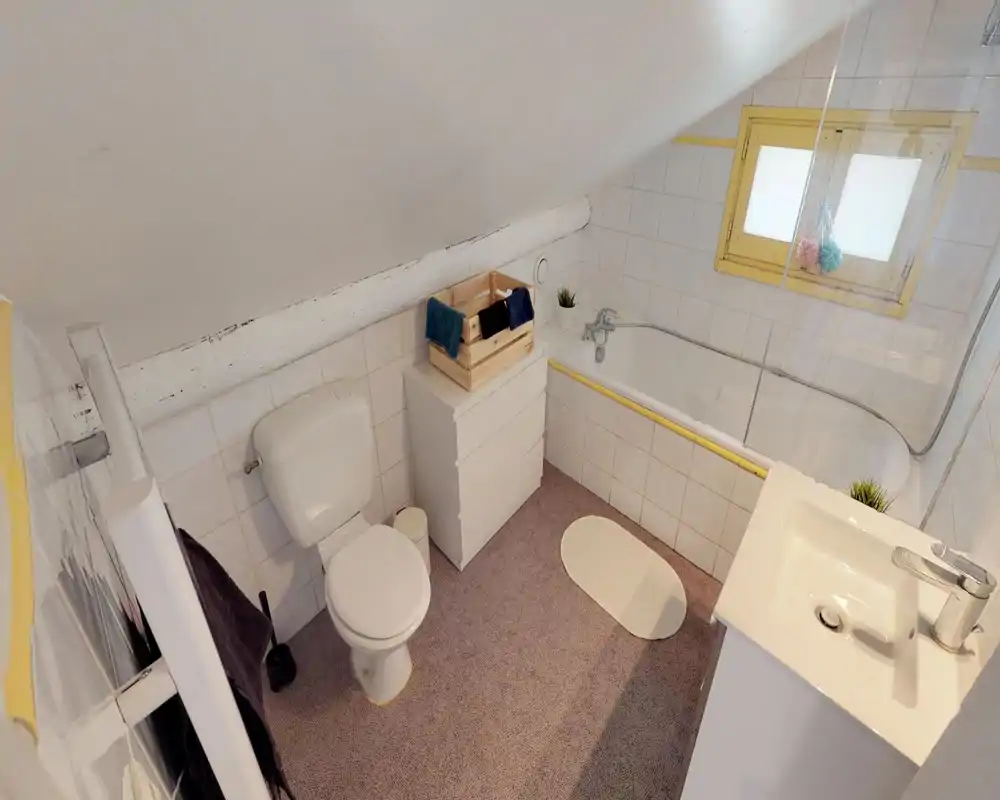 Bright attic bathroom with a shower–bath combo, toilet, sink and compact storage; white tiles and a small frosted window provide a clean, airy feel.