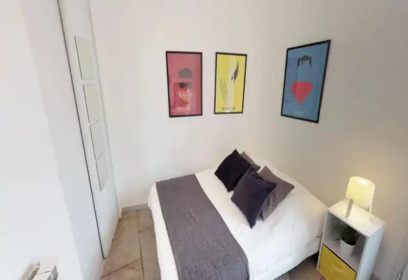 Chambre compacte et soignée avec un lit double en linge blanc, coussins décoratifs et un jeté gris. Des affiches murales minimalistes et une petite table de chevet avec lampe donnent une ambiance chaleureuse et moderne.