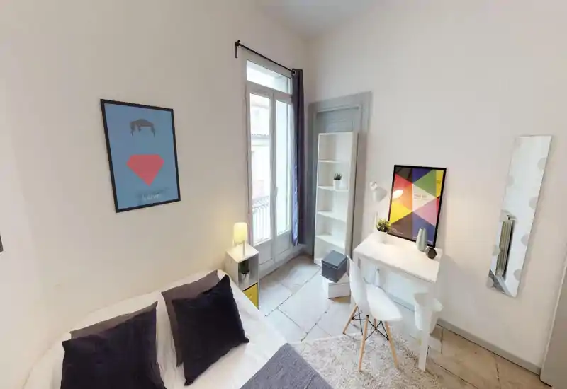 Chambre lumineuse et bien décorée avec un grand lit visible, coussins moelleux et un petit bureau près de la fenêtre. Mobilier blanc et éléments décoratifs apportent une atmosphère chaleureuse et aérée, idéale pour une annonce locative.