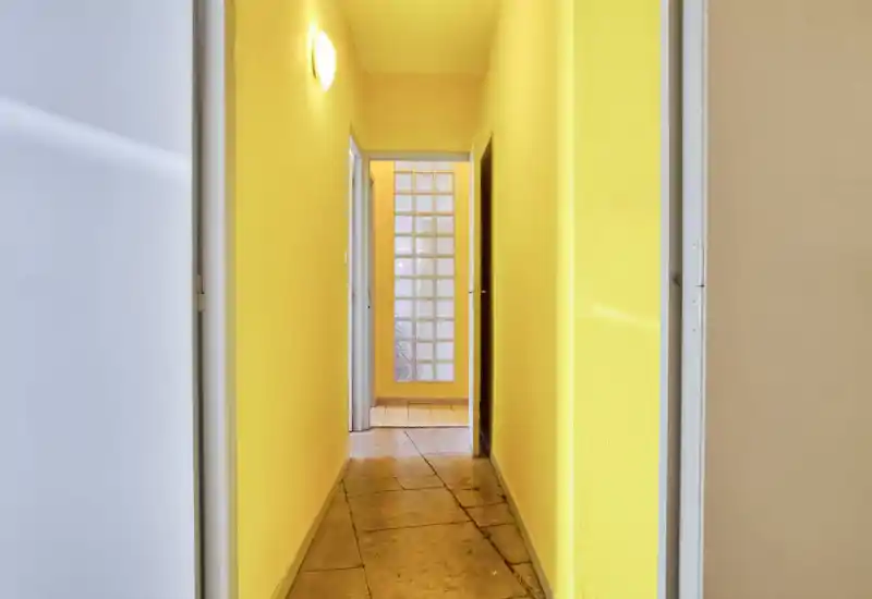 Couloir d'entrée étroit peint en jaune, bien éclairé, sol en carreaux et plusieurs portes — un passage propre et fonctionnel reliant les pièces.