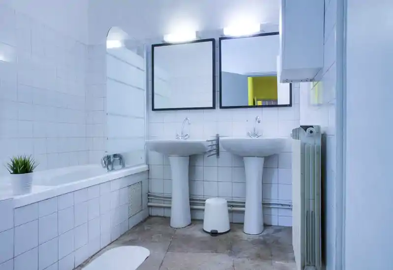 Salle de bains propre et lumineuse avec baignoire, deux vasques sur pied et miroir mural ; carrelage blanc et installations modernes épurées.