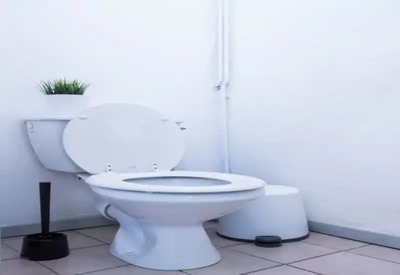 Coin de salle de bains propre et compact avec des toilettes blanches standard à siège ouvert, une petite poubelle et une brosse WC contre des murs blancs texturés.