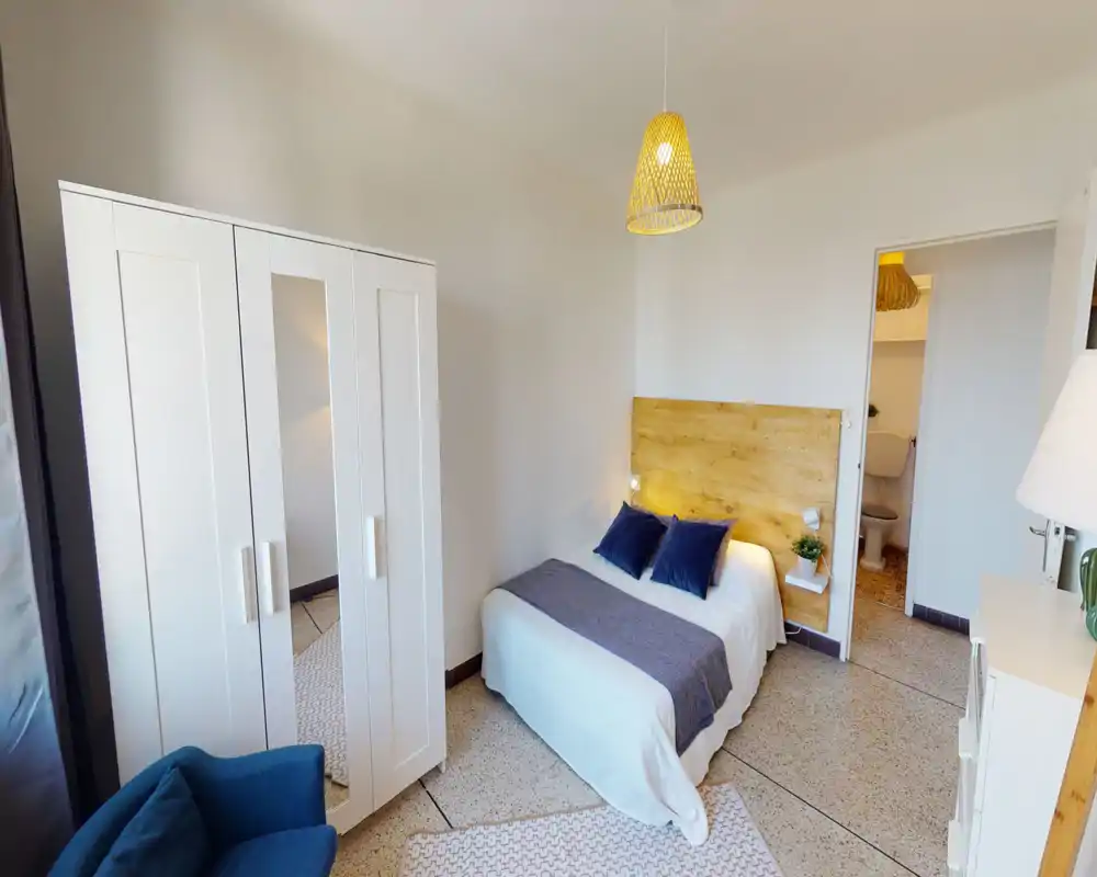 Camera da letto luminosa e accogliente con testiera in legno, letto matrimoniale con biancheria bianca e cuscini blu, armadio con specchio e poltrona comoda — perfetta per un soggiorno rilassante.