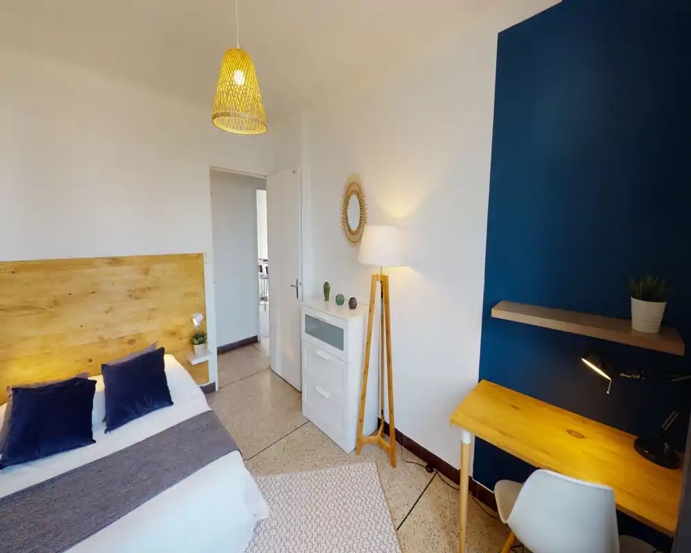 Camera da letto luminosa e curata con testiera in legno, cuscini blu navy e una scrivania su parete blu scuro. L'ambiente ha illuminazione calda, una lampada da terra e mobili contenitivi, perfetto per soggiorni confortevoli o smart working.