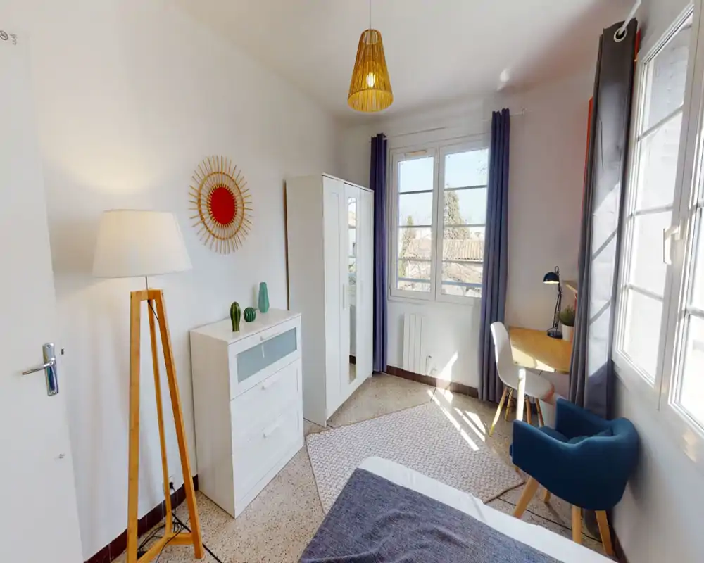 Chambre moderne et lumineuse avec lit partiellement visible, armoire et commode blanches, bureau près de la fenêtre et touches décoratives chaleureuses.