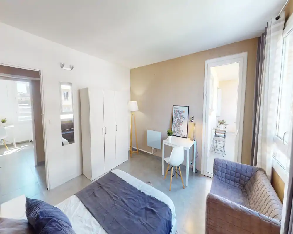 Chambre lumineuse et moderne avec lit double, armoire, petit bureau et un fauteuil confortable près de larges fenêtres laissant entrer beaucoup de lumière naturelle.